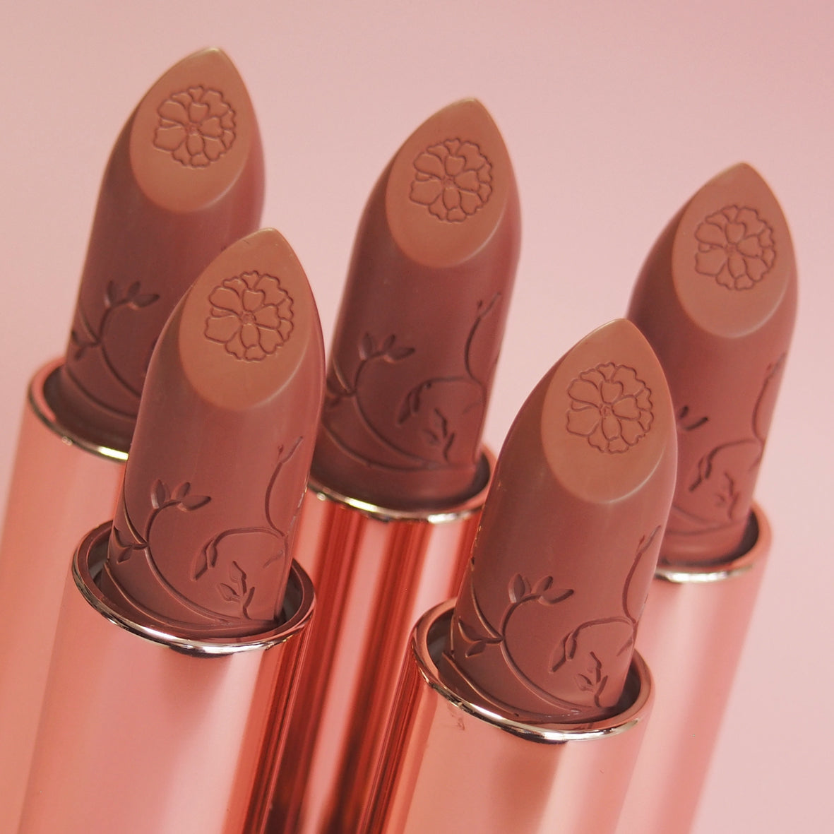 Set of five nude lipsticks with rose gold cases on a pink background