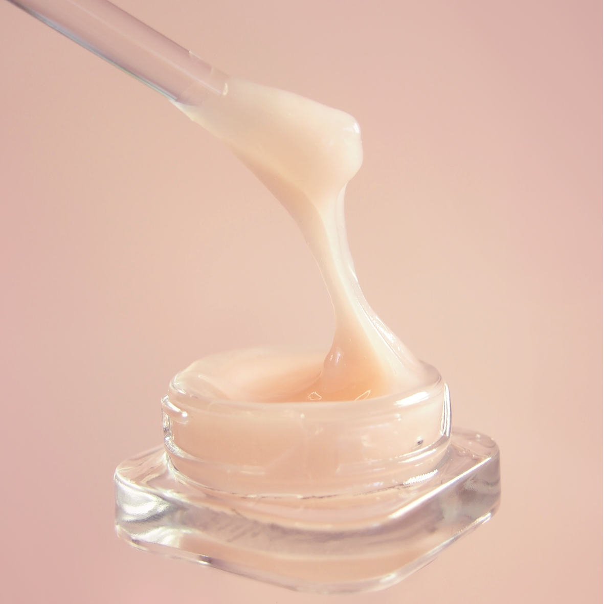 Glass jar of cream with a dropper on a pink background. 